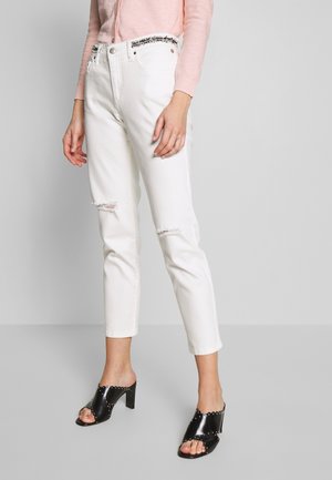 Kvinde iført hvide revnede jeans, pink langærmet top og sorte åbne hælsandaler med sølvnitter, stående foran en grå baggrund.