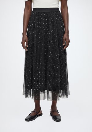 Femme portant une jupe midi plissée noire à pois blancs, associée à des chaussures plates noires et un haut blanc.
