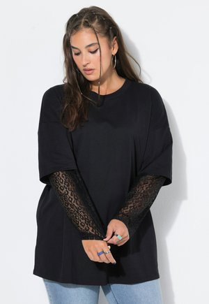 Vrouw met lang bruin haar die een zwart oversized T-shirt draagt over zwarte kanten mouwen en lichtblauwe spijkerbroek, staand tegen een witte achtergrond.