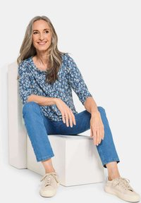 Blau gemusterte Bluse mit geometrischem Design, kombiniert mit blauen Jeans und beigen Turnschuhen. Sitzt auf einer weißen geometrischen Bank.