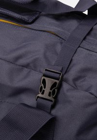 Dettaglio di una borsa in tessuto blu navy con superficie texturizzata, spallacci regolabili e fibbia in plastica nera. Include una cucitura con zip gialla a contrasto.