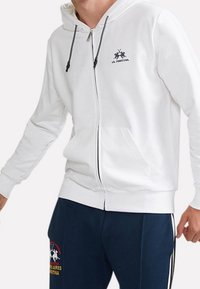 Vit zip-up hoodie tillverkad av mjukt tyg, med en ficka fram, justerbar dragsko på huvan och en logotyp tryckt på bröstet.