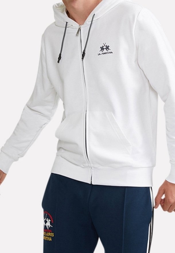 Vit zip-up hoodie tillverkad av mjukt tyg, med en ficka fram, justerbar dragsko på huvan och en logotyp tryckt på bröstet.