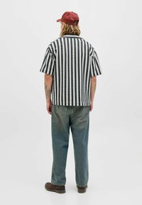 Chemise à rayures noires et blanches à manches courtes avec un col, associée à un jean léger bleu et des chaussures marron, portée à l'envers, avec des tatouages sur le bras.