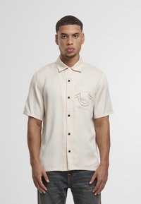 Camisa de botones de manga corta color crema con un bolsillo en el pecho y costuras negras en contraste, con botones negros y un ajuste relajado.