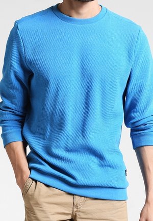 Homme portant un pull à col rond texturé bleu vif à manches longues et un pantalon beige, les mains dans les poches, sur un fond uni.