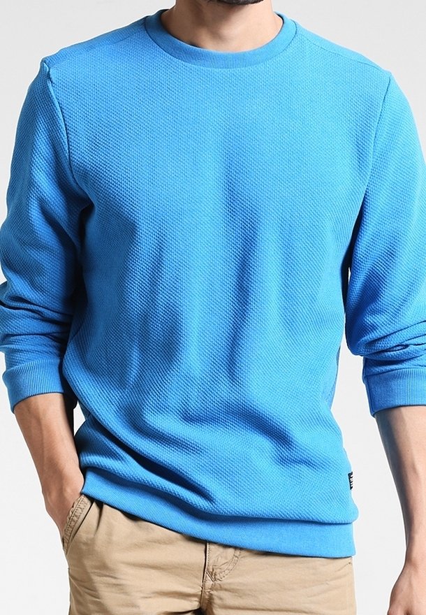 Homme portant un pull à col rond texturé bleu vif à manches longues et un pantalon beige, les mains dans les poches, sur un fond uni.