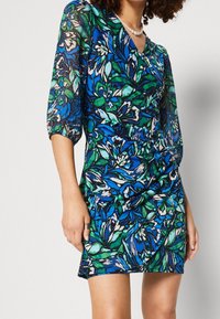 Vestido floral con un patrón vibrante en azul y verde, mangas tres cuartos y una silueta ajustada con diseño cruzado.