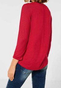 Rotes gestricktes Oberteil mit Dreiviertelärmeln und rundem Ausschnitt, ausgestattet mit einer Rückfalte. Getragen mit dunkelblauen Jeans.