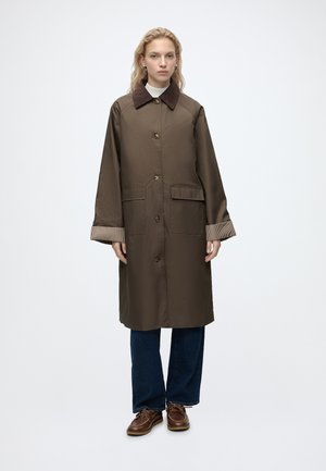 Femme portant un long manteau marron avec un col foncé, des poignets retroussés à carreaux, un jean bleu et des chaussures en cuir marron, debout devant un fond uni.