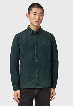 Jonge man met kort haar die een donkergroen overhemd met knopen en een zwarte broek draagt, poserend tegen een effen lichte achtergrond.