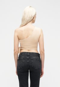 Γυναίκα με μακριά ξανθά μαλλιά που κοιτάζει προς τα πίσω, φορώντας ένα μπεζ crop top με έναν ώμο και μαύρο τζιν ψηλόμεσο, μπροστά σε ένα απλό φόντο.