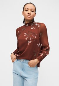 Jeune personne aux cheveux tressés portant un chemisier floral bordeaux transparent et un jean bleu clair, mains dans les poches, fond neutre.