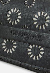 Portafoglio in pelle nera con motivi floreali ritagliati e una superficie testurizzata. Il nome del marchio "Desigual" è impressa su una striscia di pelle.