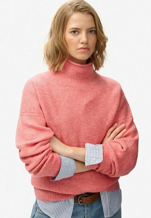 Jonge vrouw met blond haar, gekleed in een roze ribgebreide coltrui over een blauw gestreept shirt, met haar armen gekruist en een neutrale uitdrukking.