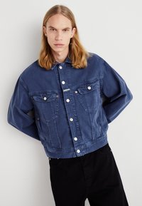 Blå denimjacka med knappstängning framtill, två bröstfickor och en avslappnad passform. Har kontrasterande sömmar och silverknappar.