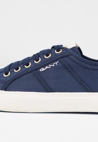 Baskets en toile bleue avec texture côtelée, œillets dorés, semelle en caoutchouc blanche et logo brodé sur le côté. Design simple et moderne.