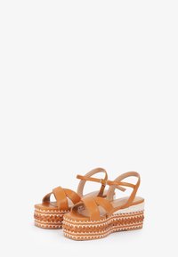 Tan leather platform sandals with cross straps and adjustable ankle strap; woven-textured sole with decorative white stitching.