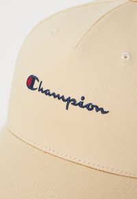 Beige színű baseballsapka textúrált anyagból, amely navy kék hímzett "Champion" logóval és egy kis piros díszítéssel rendelkezik a logón.