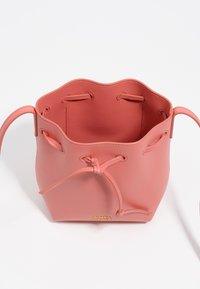 Różowa skórzana torba typu bucket z falbanką na górnej krawędzi, z zapięciem na sznurek i złotym detalem z logo. Gładka faktura z zakrzywionymi uchwytami.