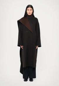 Abrigo largo negro con un collar marrón asimétrico, diseño oversize, mangas anchas y cierre de un solo botón. Textura suave.