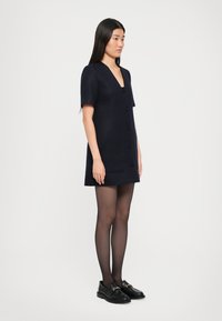 Tengerészkék, rövid ujjú ruha V-kivágással és dupla gombos dizájnnal, texturált anyagból készült. Fekete loaferekkel és áttetsző harisnyával viselve.