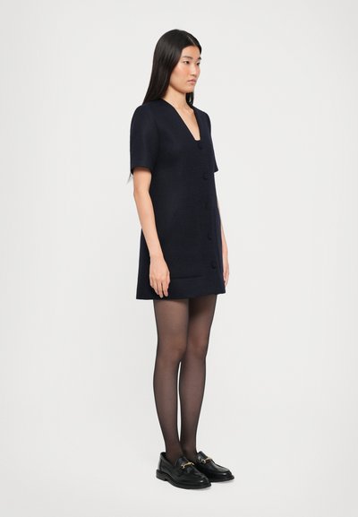 Rochie bleumarin cu mâneci scurte, decolteu în V și design cu nasturi dubli, realizată dintr-un material texturat. Asortată cu loafers negri și ciorapi transparenți.