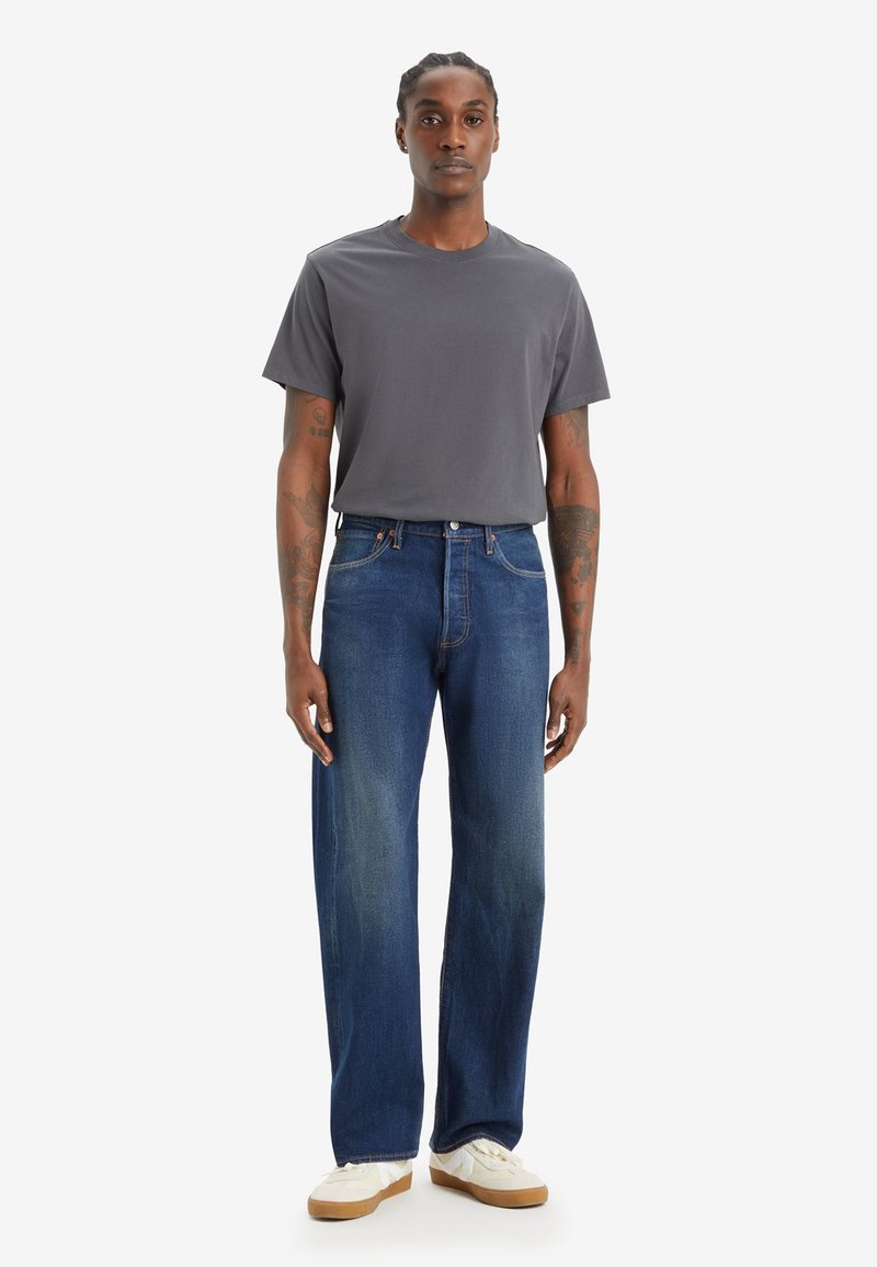 T-shirt grigia abbinata a jeans in denim blu. I jeans presentano una leggera scoloritura, un design a gamba dritta e sono indossati con sneakers bianche.