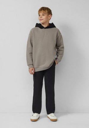 Ragazzo in piedi in modo informale, indossa un felpa grigia con cappuccio nero, pantaloni neri e sneakers bianche, su uno sfondo chiaro e semplice.