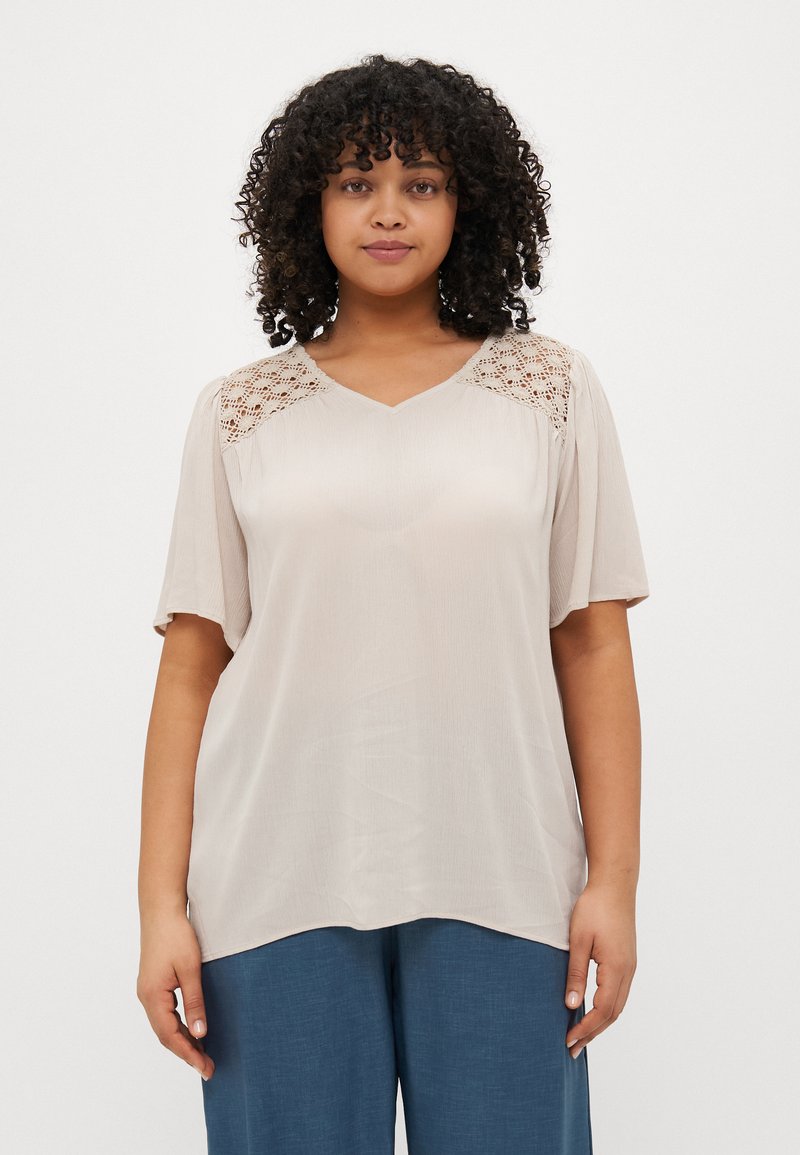 Beige bluse med V-hals, korte ermer og intrikate blonder på skuldrene. Mykt, flytende stoff med en avslappet passform.