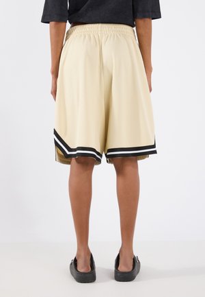 Person trägt beige knielange Shorts mit schwarzen und weißen Streifen am Saum, schwarzes Hemd und schwarze Slipper, steht mit dem Rücken zur Kamera.