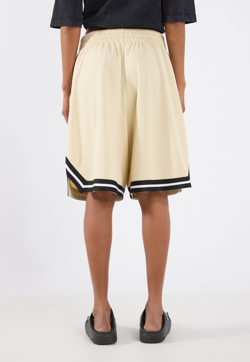 Personne portant un short beige mi-genou avec des rayures noires et blanches à l’ourlet, une chemise noire et des chaussures noires sans lacets, debout de dos.