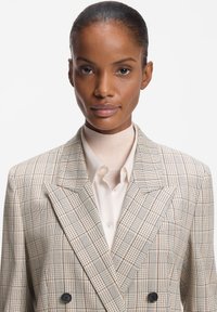 Dobbeltradet blazer i et lyst beige ternet mønster med sorte og røde accenter. Tekstureret stof med hakkede revers og sorte knapper.