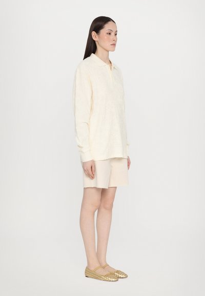 Polo a maniche lunghe crema con un motivo tessutato, abbinato a pantaloni corti beige. Scarpe slip-on in oro intrecciate completano l'outfit.
