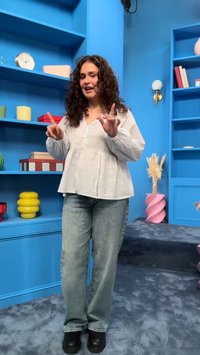 Žena s kovrčavom kosom u bijeloj bluzi i trapericama gestikulira objema rukama u šarenoj sobi s plavim policama i ukrasnim predmetima.