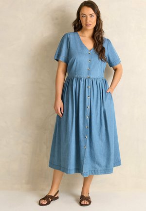 Frau mit langen braunen Haaren, die ein blaues, knöpfbares Midikleid mit kurzen Ärmeln und braunen Sandalen trägt, steht mit einer Hand in der Tasche.