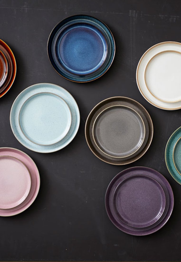 Set mit neun Keramiktellern in verschiedenen Farben: Rost, Dunkelblau, Mintgrün, Hellrosa, Dunkelgrau, Weiß und Lila, mit strukturierten Oberflächen und Randakzenten.
