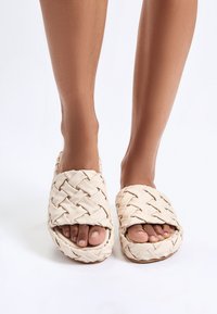 Geweven, beige slide-sandalen met een dikke zool. Open teen ontwerp onthult de tenen. Heeft een gestructureerd, gevlochten patroon op de bovenkant.