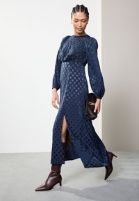 Marineblaues, langärmeliges Kleid mit Punktmuster, seitlichem Schlitz und tailliertem Schnitt, kombiniert mit einer schwarzen, strukturierten Clutch und braunen, spitzen Stiefeln.
