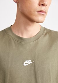 Olīvu zaļa kokvilnas t-krekls ar ribotu apļa kakla izgriezumu un baltu Nike logo izšuvumu uz krūtīm. Gluda tekstūra, minimālisma dizains.