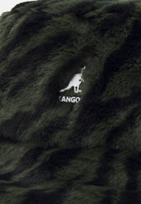 Texture en fausse fourrure verte avec un motif de fines rayures noires. Est doté d'un logo Kangol blanc brodé à l'avant. Matériau doux et pelucheux.