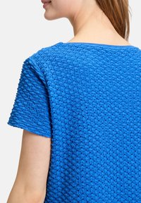 Blaues, strukturiertes T-Shirt mit kurzen Ärmeln, das ein gestricktes Muster mit kleinen Löchern aufweist. Nahtloser Ausschnitt und lockerer Sitz für höchsten Komfort.