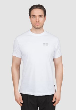 Hombre con camiseta blanca con texto negro "KARMA KOMMT. KARMA REGELT." en el pecho izquierdo, combinado con pantalones negros.