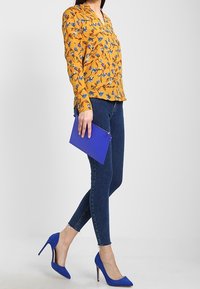 Blusa amarilla con flores, jeans ajustados azules, bolso de mano azul y tacones altos azules. La blusa tiene un escote en V y mangas largas; el denim es ajustado.