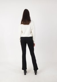 Witte, gestructureerde trui gecombineerd met strakke zwarte flared broeken en hoge zwarte schoenen. Gezicht vanaf de achterkant, tegen een effen achtergrond.