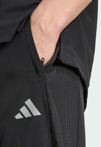 Shorts de sport noirs avec un panneau texturé, dotés d'un logo Adidas argenté sur la jambe inférieure et d'une poche latérale zippée.