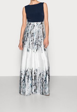 Kobieta w granatowym topie bez rękawów oraz białej spódnicy do ziemi z wysokim stanem, ozdobionej czarnymi i szarymi pociągnięciami pędzla.