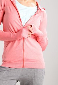 Person in einem hellrosa Zip-Hoodie mit Daumenlöchern, einem weißen Shirt darunter und grauen Hosen, der vor einem schlichten Hintergrund steht.