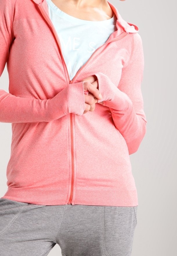 Person in einem hellrosa Zip-Hoodie mit Daumenlöchern, einem weißen Shirt darunter und grauen Hosen, der vor einem schlichten Hintergrund steht.