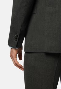 Veste de costume gris foncé avec un tissu texturé, comportant un poignet à trois boutons et une coupe nette à l'ourlet, assortie de pantalons gris.